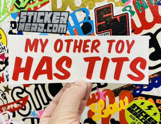 Sticker with text 'MY OTHER TOY HAS TITS' held over a colorful sticker background.
