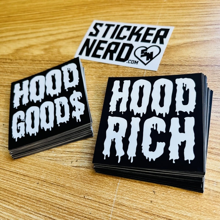 Stacks of 'Hood Goods' and 'Hood Rich' stickers with a 'Sticker Nerd' sticker on a wooden surface.