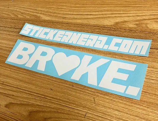 Two light blue rectangular sticker decals on a wooden surface. The top sticker reads Broke.