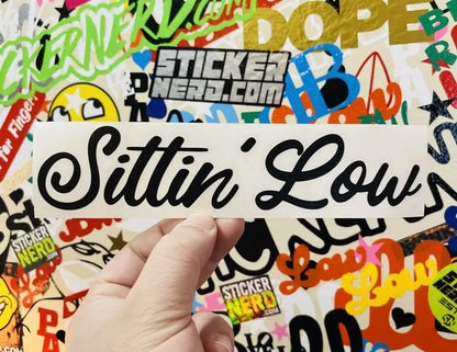 A hand holds a white rectangular sticker with the phrase 'Sittin' Low' in black cursive script, set against a background covered in colorful stickers, including visible 'Sticker Nerd' branding.