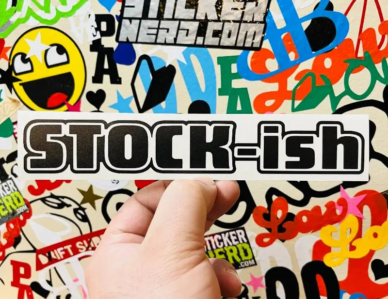 A hand holds a white rectangular sticker with the text 'STOCK-ish' in bold black letters with a white outline. The background is a dense collage of colorful stickers and graphics, including 'STICKER NERD.