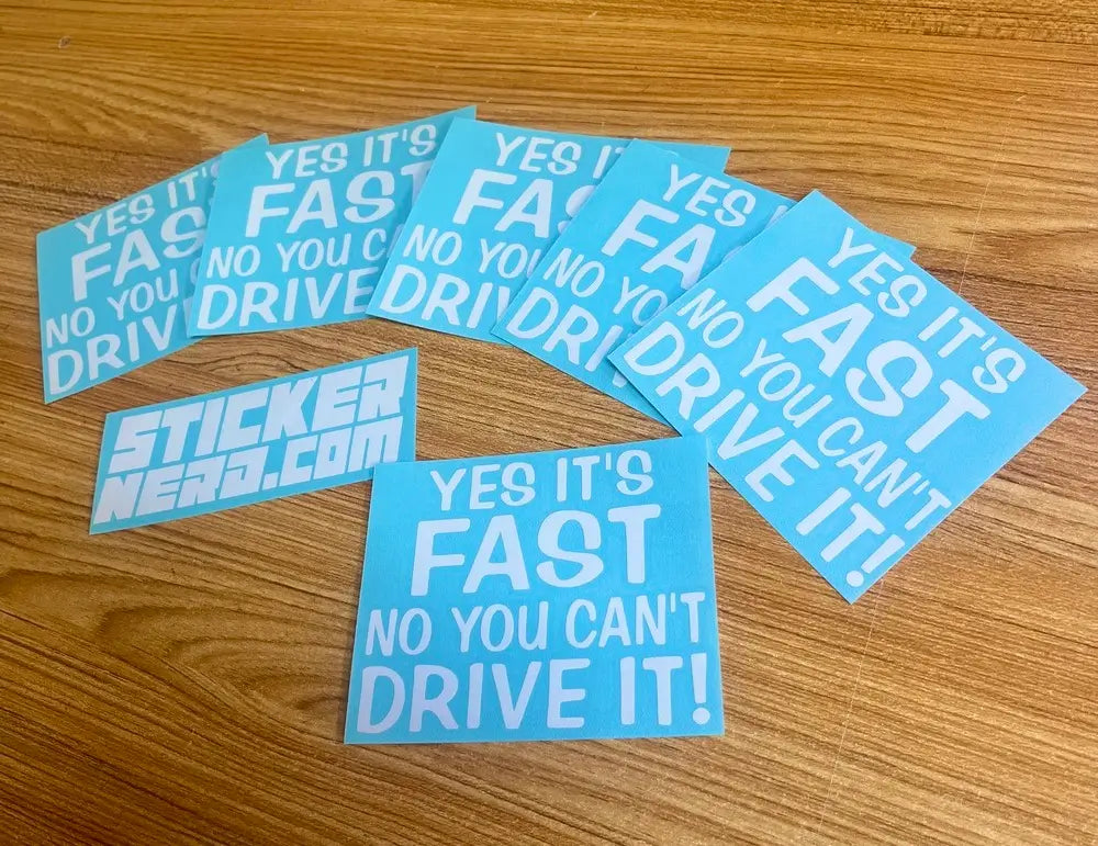 A set of light blue vinyl stickers with white text reading 'YES IT'S FAST NO YOU CAN'T DRIVE IT!' and one sticker reading 'STICKER NERD.COM', arranged on a wooden tabletop.